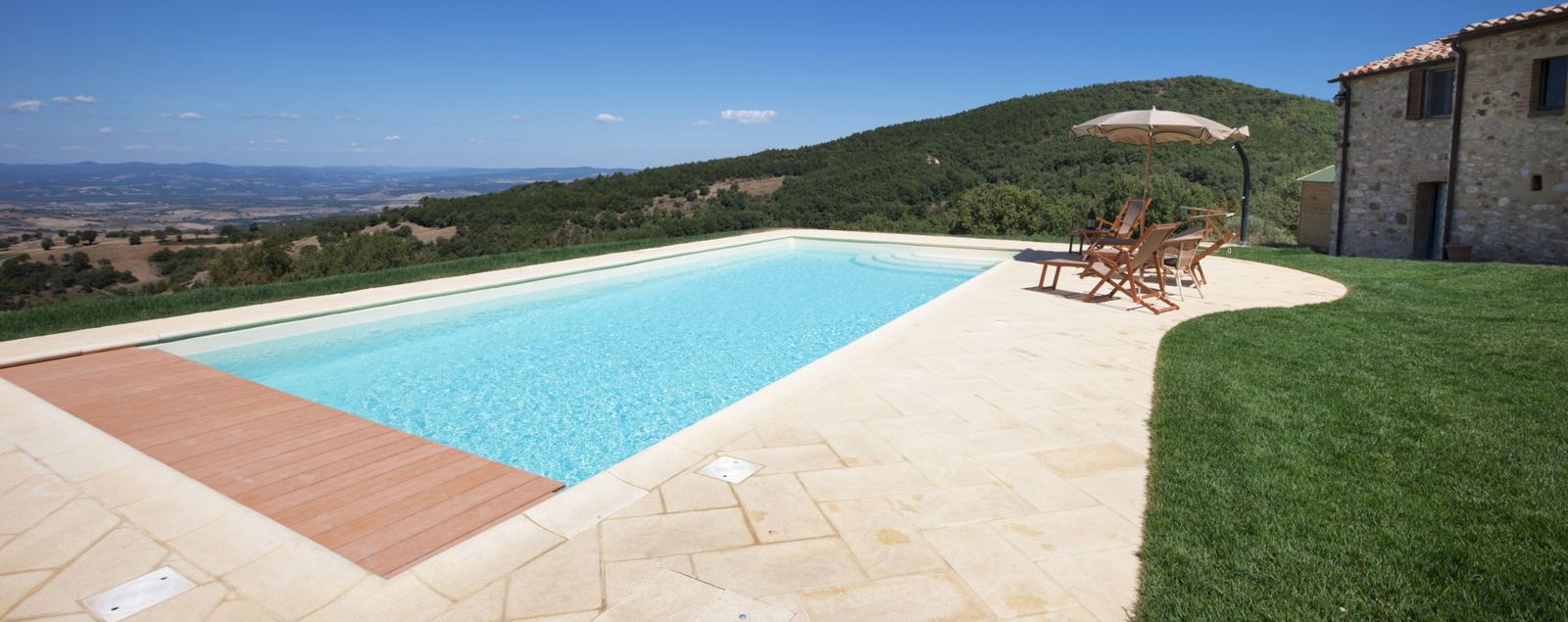 Piscina con solarium in pietra e deck in legno davanti a un casale